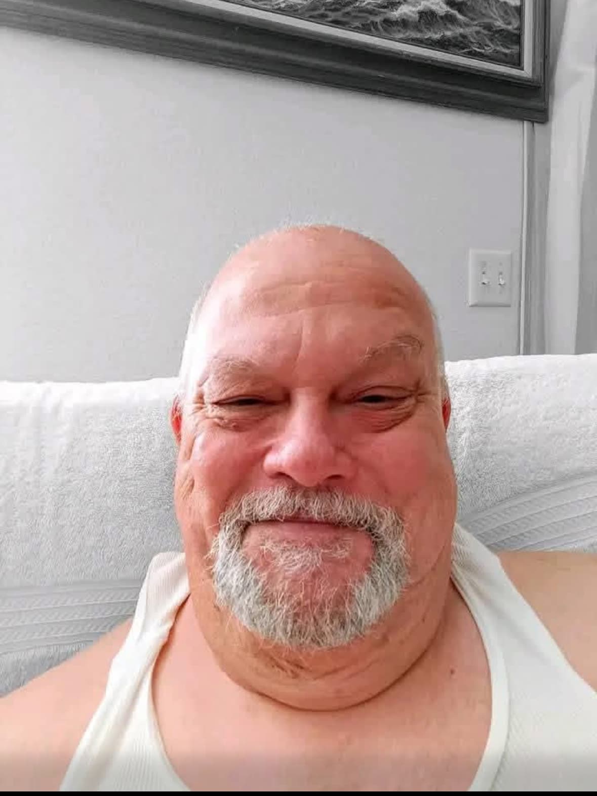 Smiling bald man with a grey goatee wearing a white tank top against a wall.